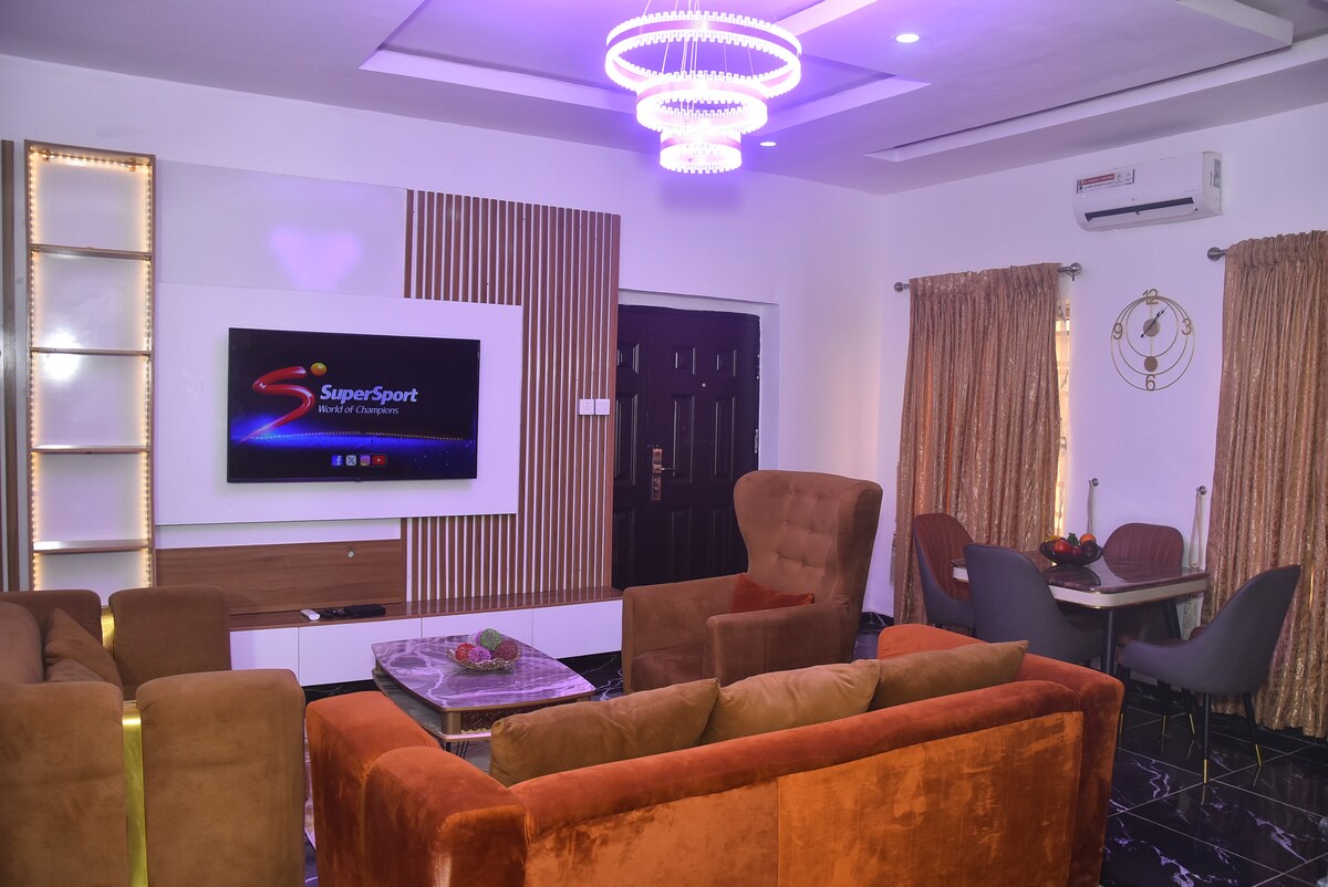 A modern living area features a plush brown sofa and two comfortable armchairs, inviting relaxation. A large flat-screen TV is mounted on the wall, surrounded by stylish decor. Elegant curtains frame the windows, and a dining area with a table and chairs is visible in the background.