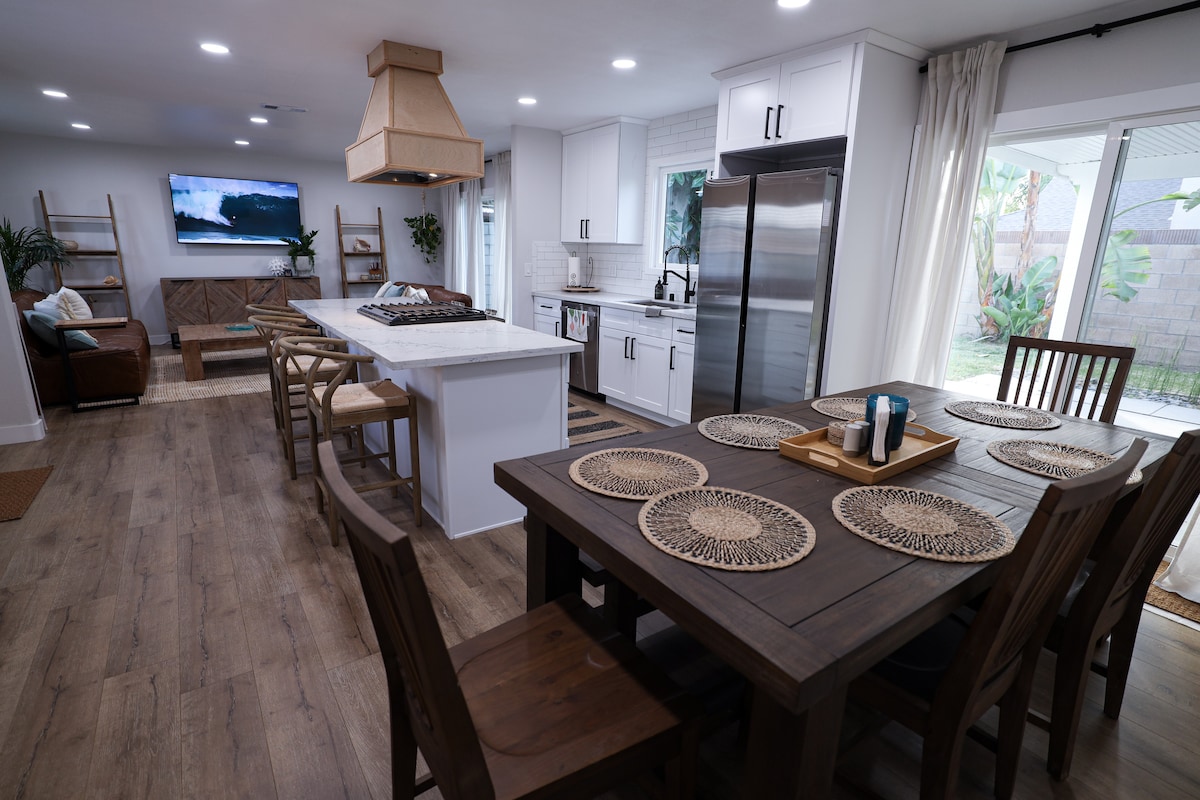 A spacious kitchen and dining area is shown, featuring a large wooden table set for six. Modern appliances and a central island are present in the kitchen, with a stylish range hood overhead. Natural light enters through glass doors, illuminating the open space.