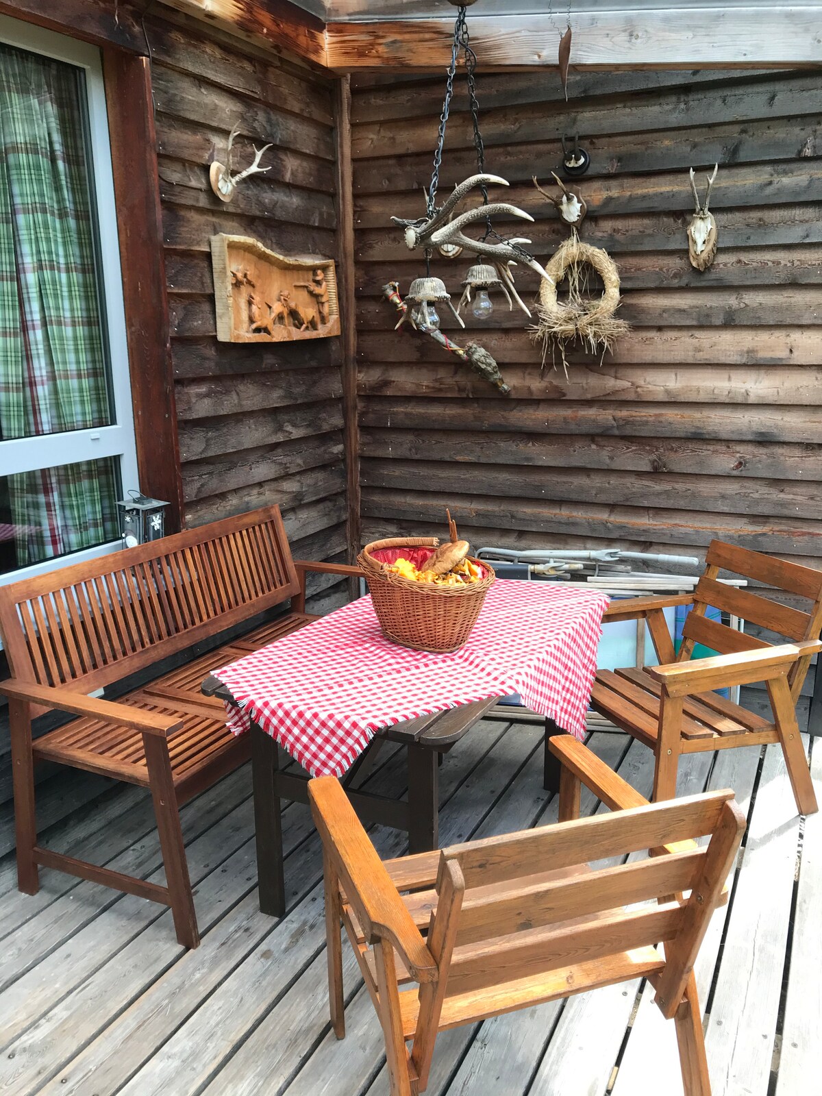 A wooden terrace is furnished with a checked tablecloth covering a small table surrounded by four wooden chairs. Decorative elements, including mounted antlers, are displayed on the walls, contributing to the rustic charm of the space.
