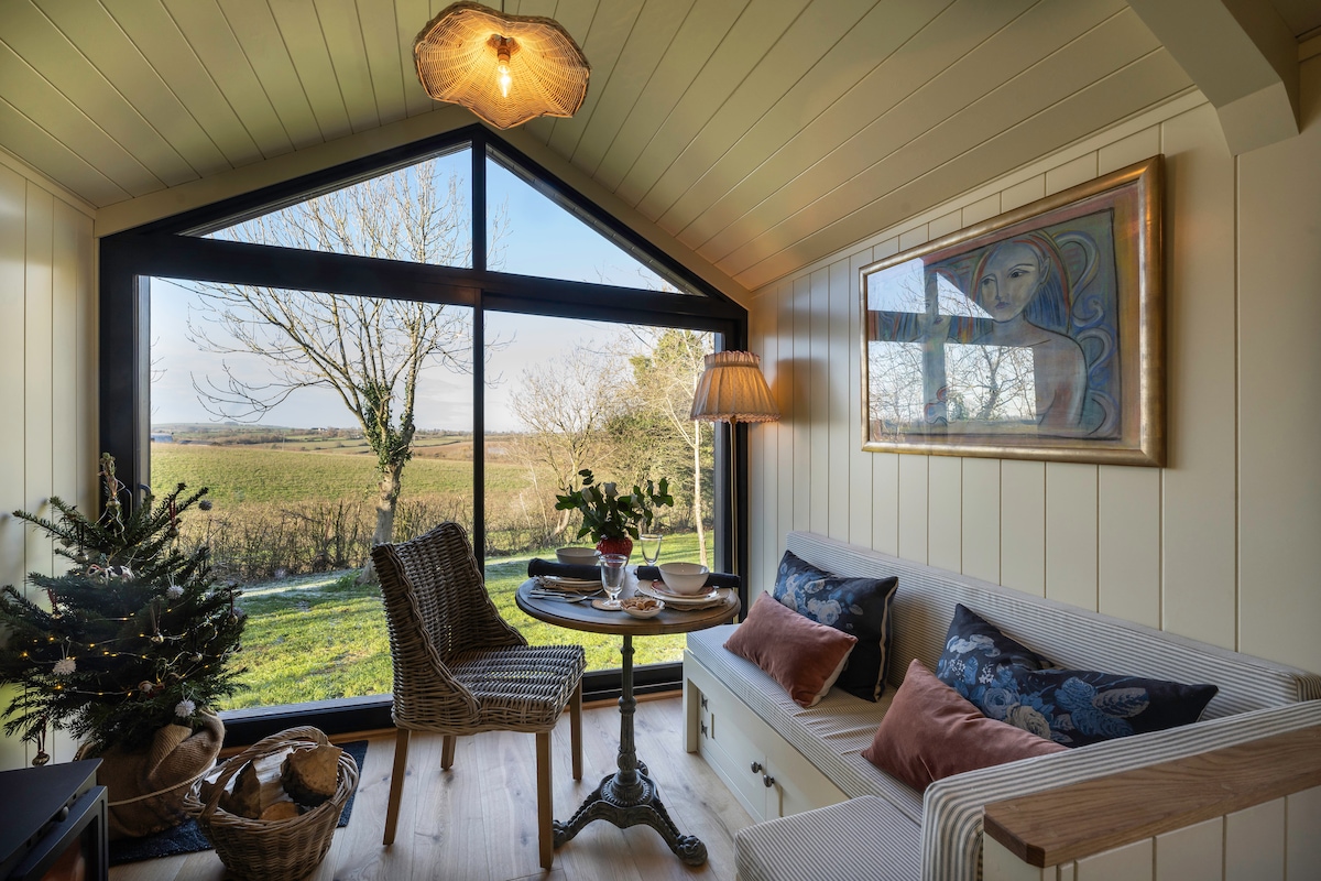 A bright sitting area features large windows that frame the outdoor landscape of rolling fields. A comfortable sofa is accompanied by decorative cushions and a small round table, set for two with a vase of flowers. A softly lit lamp enhances the inviting ambiance.