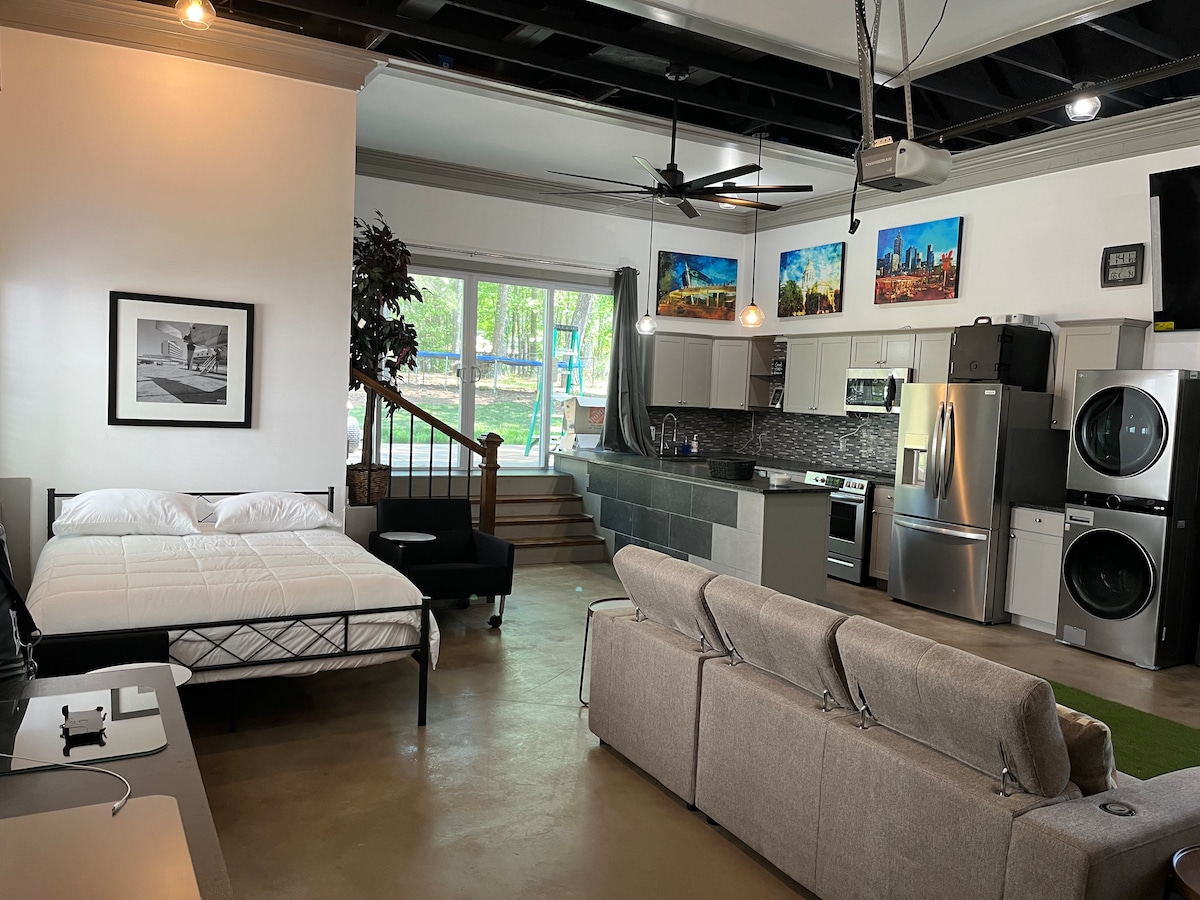 A studio-style living room is showcased, featuring a spacious bed positioned near a wall, with a comfortable sofa facing a large kitchen area. Modern appliances, including a refrigerator and washing machine, are visible. Large windows provide natural light and views of the outdoors.