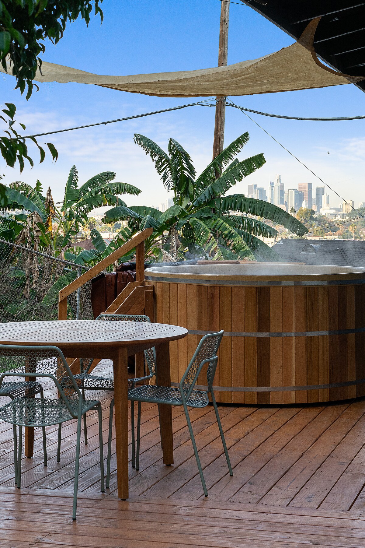 A wooden deck features a round Japanese cedar hot tub, steam rising from the water. A circular table with four chairs complements the outdoor space. Lush green banana trees frame the view, which includes a skyline of Downtown LA in the background.