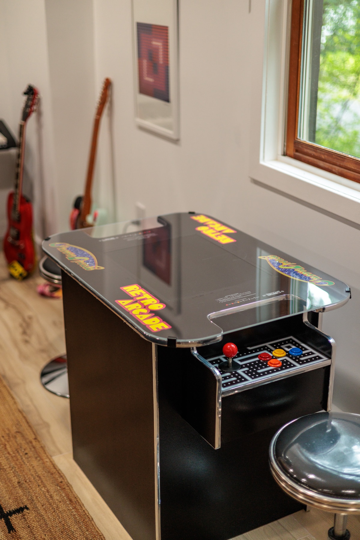A vintage style arcade game table is positioned against a white wall, featuring colorful graphics on its surface and control panel. A chrome stool is situated nearby, complementing the sleek design. In the background, two guitars are displayed, adding to the room's entertainment theme.