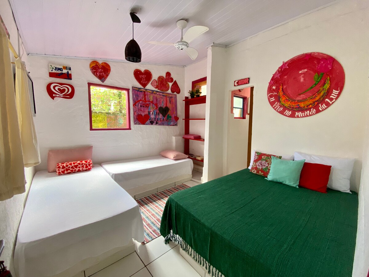 The guest suite features a spacious bed adorned with colorful cushions in tones of green, pink, and white. A second bed is positioned nearby, with soft linens. Walls are decorated with vibrant local artwork, creating a warm and inviting atmosphere. Natural light enters through windows with simple treatments.