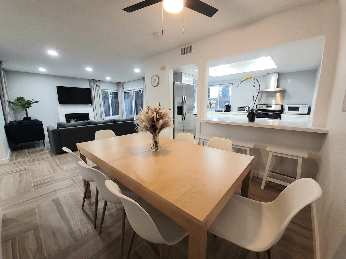 A spacious dining area features a light wooden table surrounded by white chairs. A simple vase of dried flowers adds a decorative touch. The open layout allows visibility to a well-equipped kitchen and a cozy living room area, illuminated by natural light.