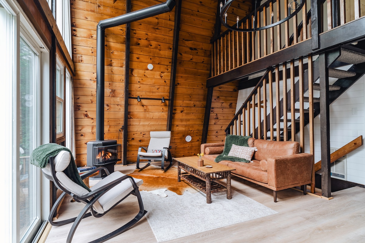 The living area features a mix of rustic charm and modern comfort, with a brown leather sofa and a cozy rocking chair on a light rug. A stylish coffee table sits in front of a gas fireplace, accompanied by wood paneling and large windows that allow natural light to fill the space.