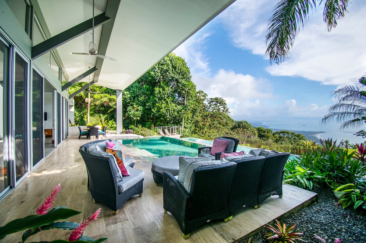 A spacious outdoor area is presented with comfortable seating arranged around a circular table. The swimming pool reflects the clear sky, surrounded by vibrant tropical foliage. Large glass doors provide a seamless connection to the interior, inviting natural light and views of the surrounding landscape.