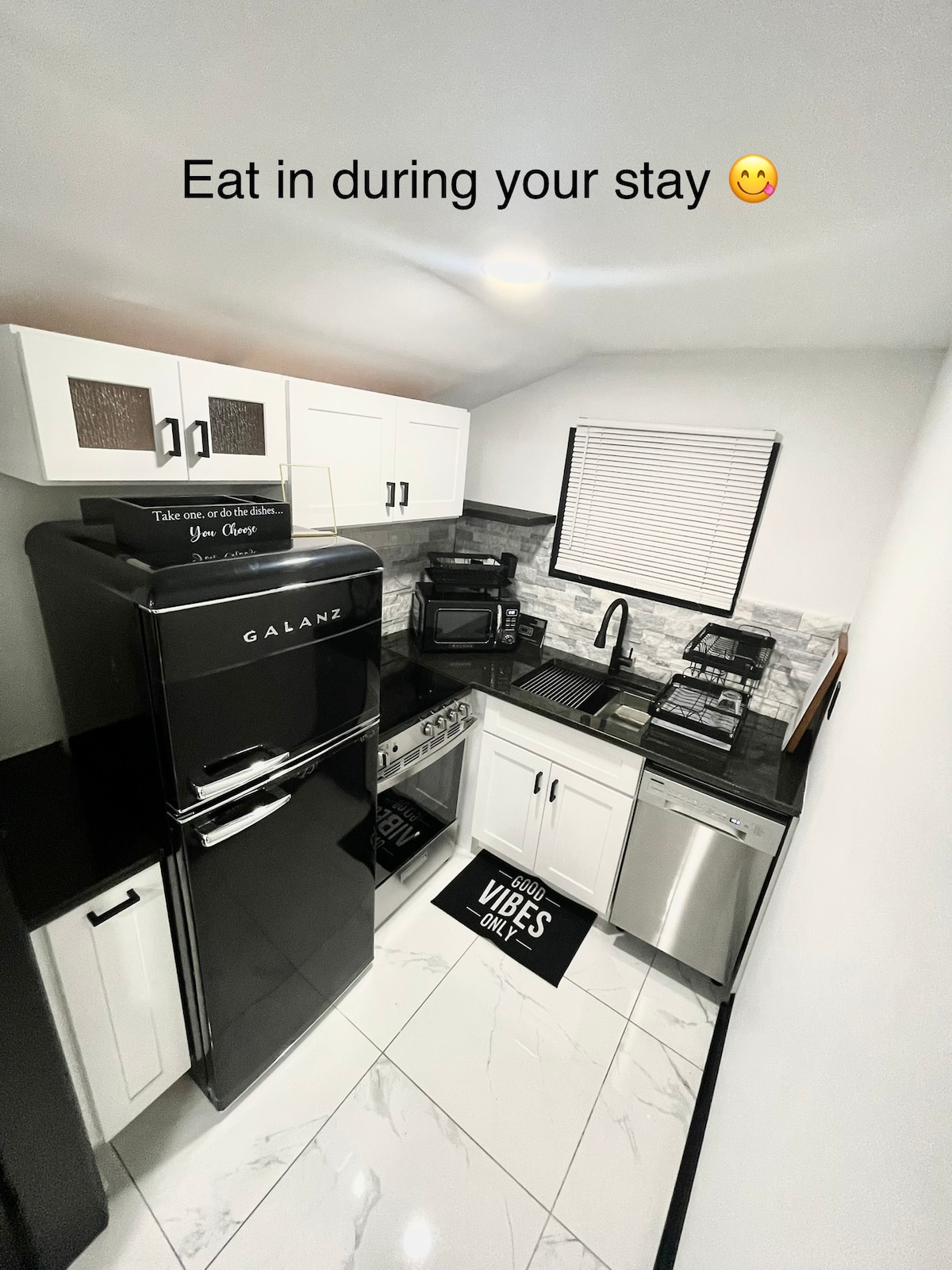 The kitchen features modern appliances, including a sleek black refrigerator, stainless steel dishwasher, and oven. Countertops in a dark hue provide a stylish space for meal preparation. A large sink is situated next to the window, with kitchen utensils and cookware neatly arranged.
