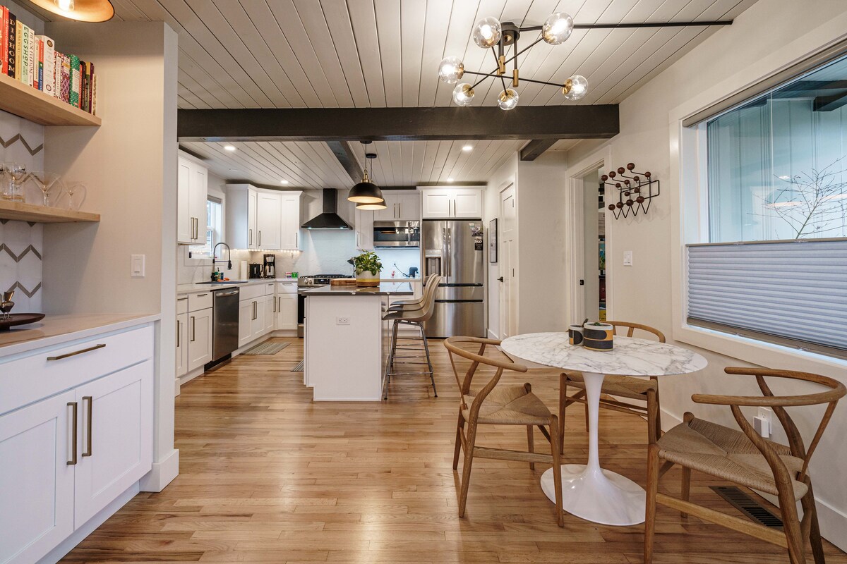 An open kitchen and dining area feature clean lines and modern finishes. A circular marble table is surrounded by light wooden chairs, while a center island showcases additional seating. Bright windows allow natural light to fill the space, complemented by warm wood flooring.