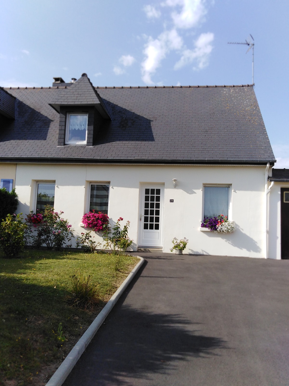 A charming house is depicted, featuring a peaked roof and white exterior. Flower boxes with vibrant blooms are placed under the windows. A paved driveway leads up to the entrance, surrounded by well-maintained greenery.