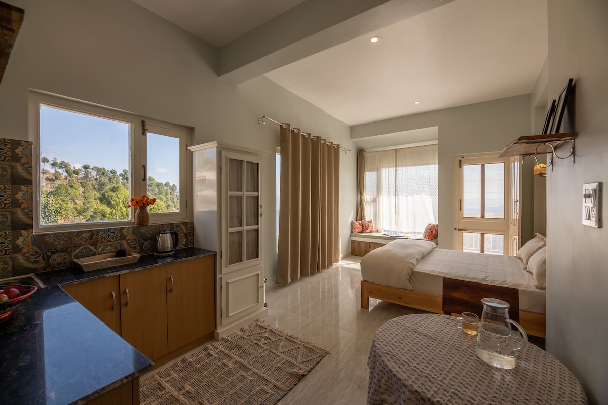 A bright and airy room features a spacious bed near large windows, offering views of surrounding hills. A kitchenette with wooden cabinets is visible, complemented by a round table with drinks. Natural light floods the space, highlighting soft furnishings and decor.