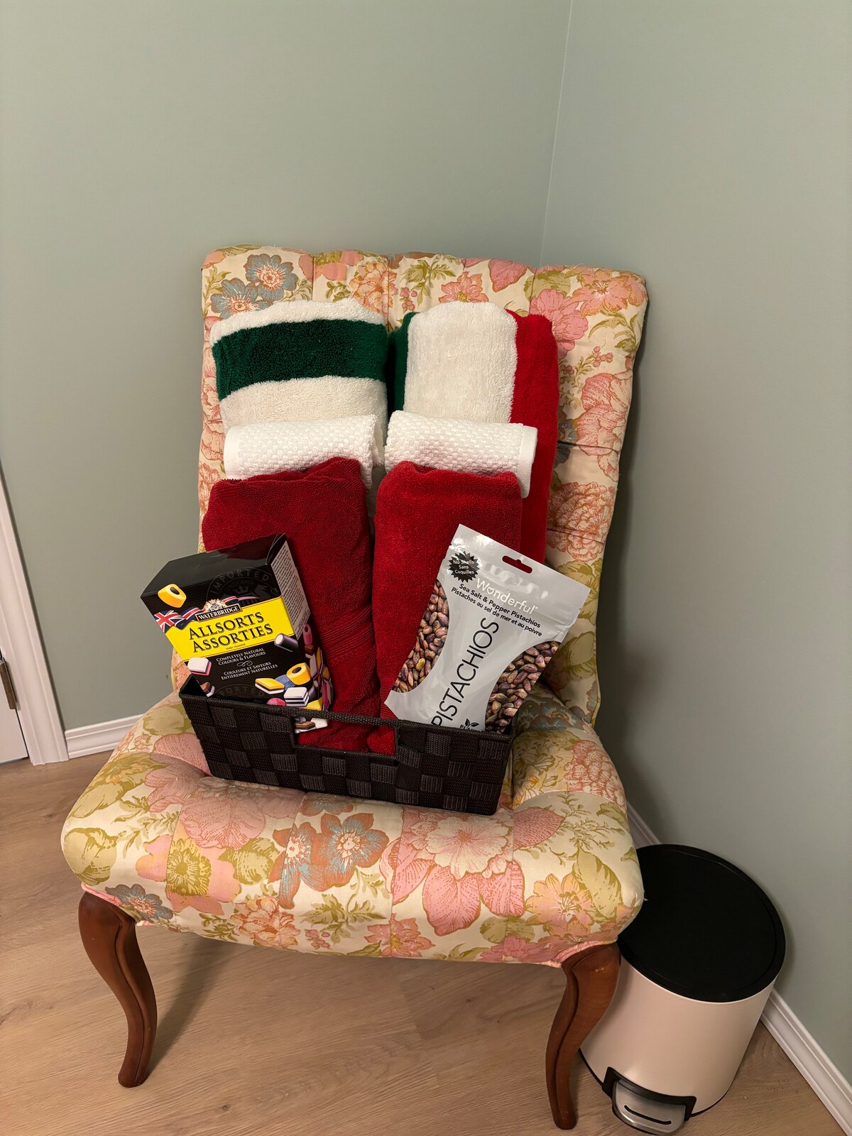 A decorative chair features a floral patterned upholstery. A basket holds neatly stacked towels in red and green, accompanied by snacks packaged in colorful bags. A small trash bin is positioned beside the chair against a light green wall.