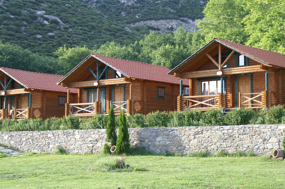 Three wooden cabins are set against a backdrop of lush greenery and a hillside. Each cabin features a front porch with railings, large windows, and a sloped roof with red tiles, providing a warm and rustic appearance.