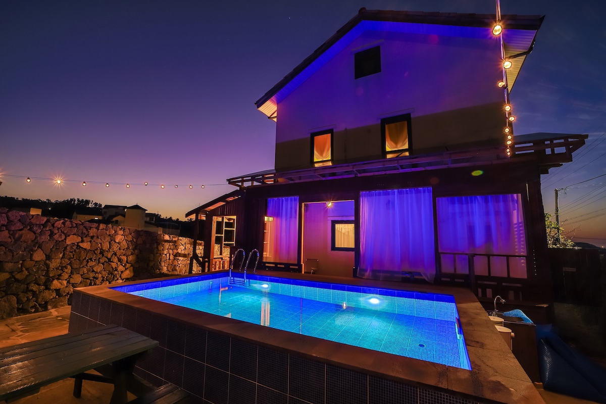 The exterior of a two-story villa is illuminated by soft lighting, highlighting the swimming pool filled with inviting blue water. The surrounding area features stone walls and large windows covered by sheer curtains, creating a tranquil setting at dusk.