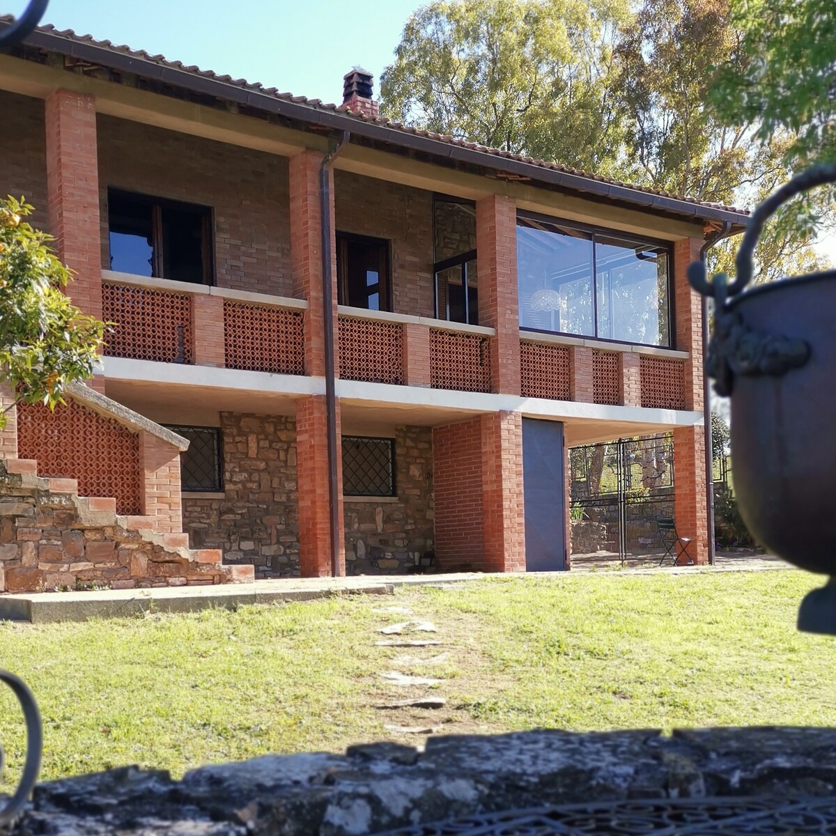 The exterior of a two-story house is visible, showcasing a blend of brick and stone construction. A series of large windows offers views of the surroundings. A covered balcony is seen above, supported by red-brick columns, with steps leading down to a grassy area.