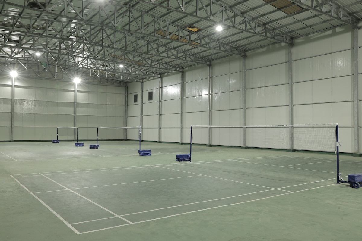 An indoor sports facility is shown, featuring a spacious court for badminton. Multiple nets are set up on either side of the court, with blue bases supporting them. The area is well-lit with overhead lighting, and high walls are lined with windows at the top.