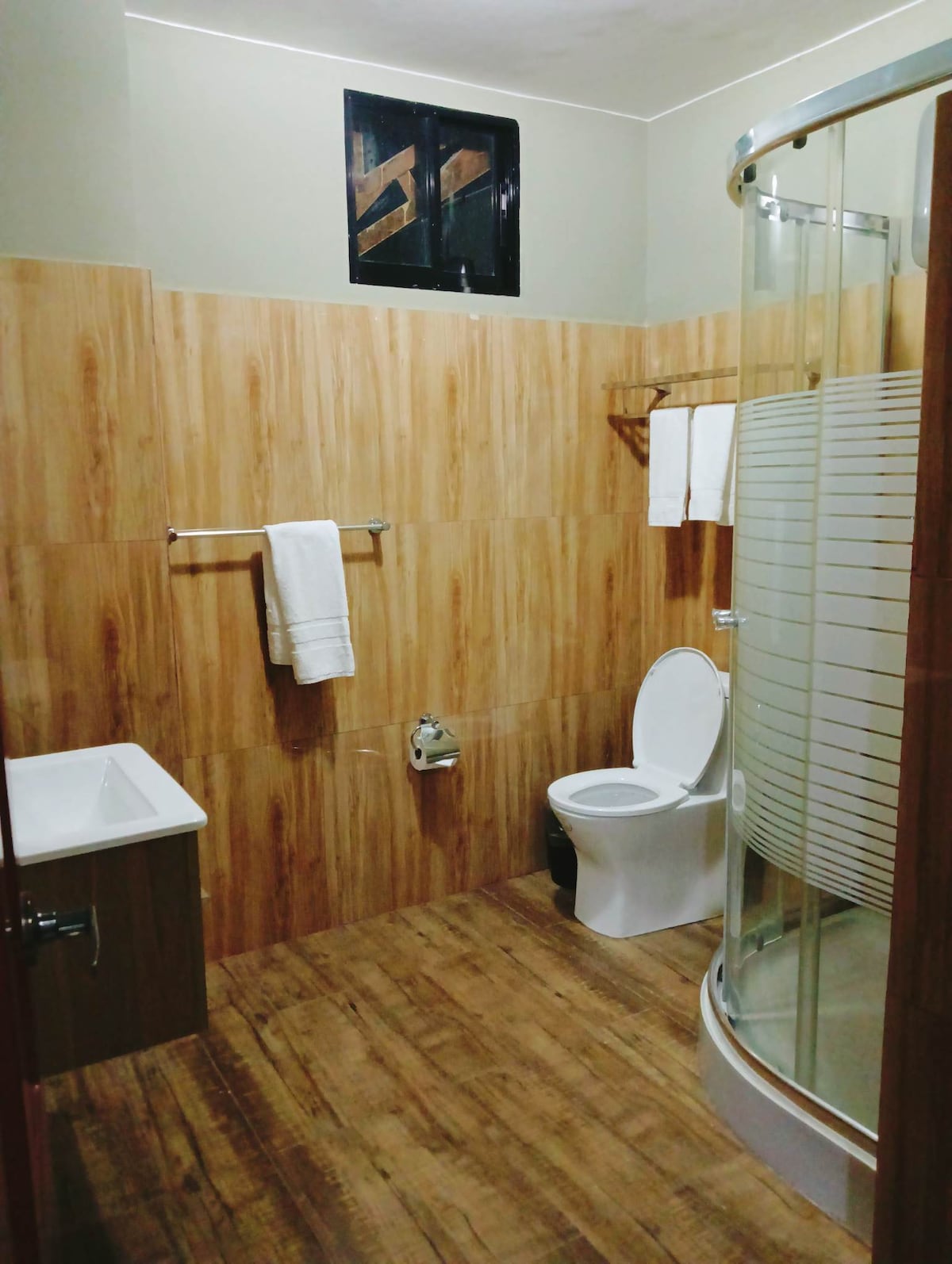 A modern bathroom features wooden paneling on the walls and floor, creating a clean aesthetic. A circular shower enclosure is visible, along with a white toilet and a washbasin. Fresh white towels are neatly hung by the wall, complementing the space.