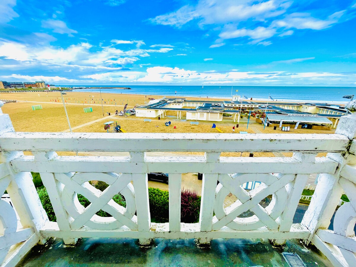 A balcony railing frames a panoramic view of the beach and sea. The sandy shore is visible, along with recreational facilities and vibrant sky above. Light clouds create a dynamic backdrop against the blue horizon, enhancing the coastal atmosphere.