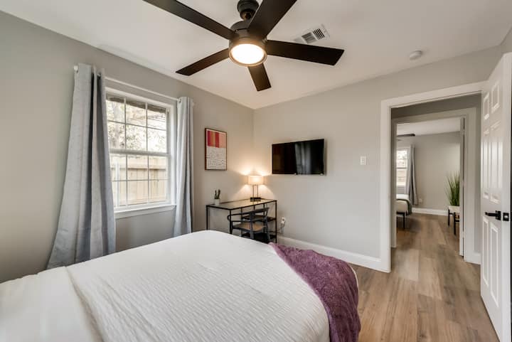  Queen bed with white bedding and purple throw. Ceiling fan, wall-mounted TV, and a small table under it. A window with curtains offers natural light.