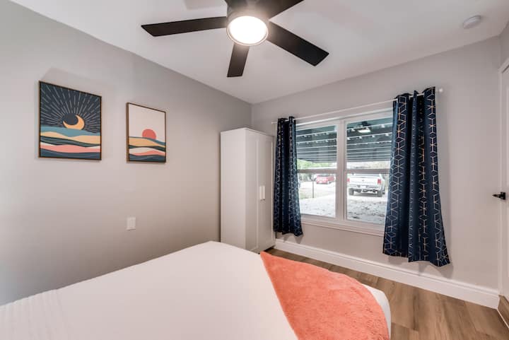 Queen bed with orange throw and sunset-themed wall art. Ceiling fan, wardrobe, and window with dark curtains complete the layout.