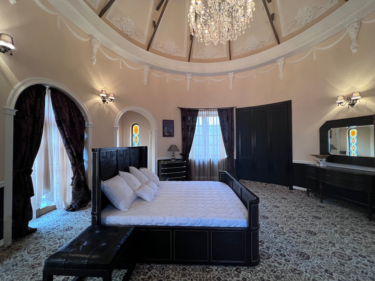 A spacious circular bedroom features a large bed with white bedding at its center. Elegant chandeliers hang overhead, while tall windows dressed in dark curtains allow natural light to filter in. A dark wooden dresser and mirror are positioned against one wall, complementing the ornate decor.