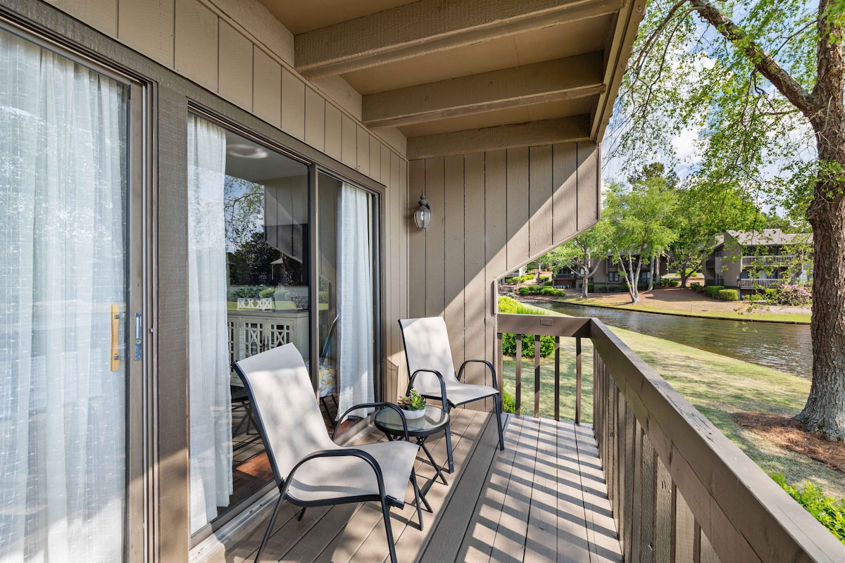 An outdoor patio is revealed, featuring two light-colored chairs positioned on a wooden deck. Sunlight casts gentle shadows on the floor, with a view of Lake Pinehurst visible beyond. Lush greenery frames the scene, enhancing the serene atmosphere.