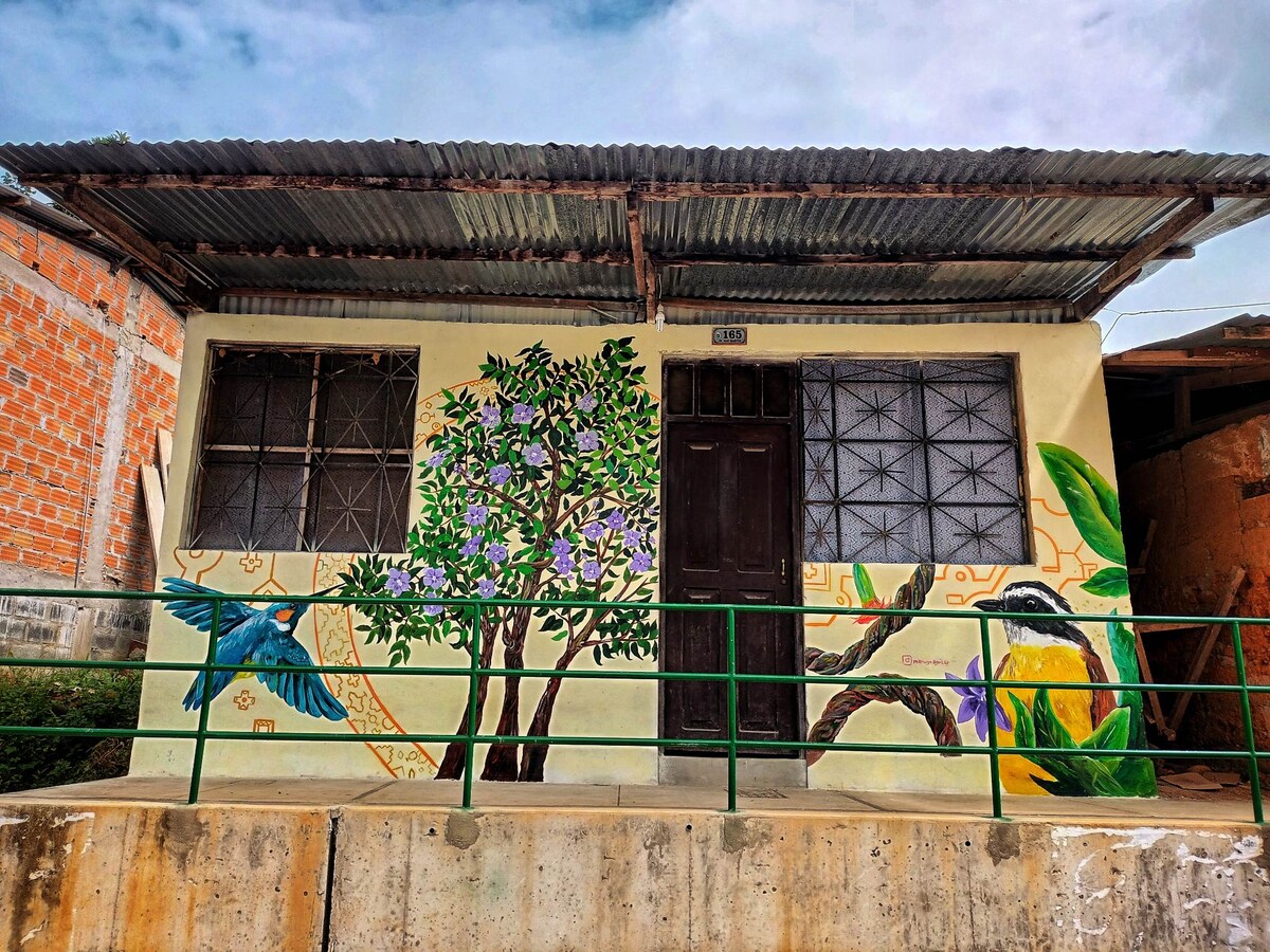 A small house is depicted with a vibrant mural featuring a tree and colorful birds. The structure has two windows with metal grilles and a brown door. A green railing runs along the front, contrasting with the natural surroundings.