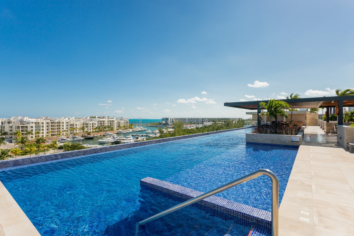 A rooftop infinity pool reflects the clear blue sky, offering stunning views of the marina and surrounding buildings. Loungers and shaded seating areas are positioned nearby, creating inviting spaces for relaxation and enjoyment. Lush greenery is visible along the pool's edge.