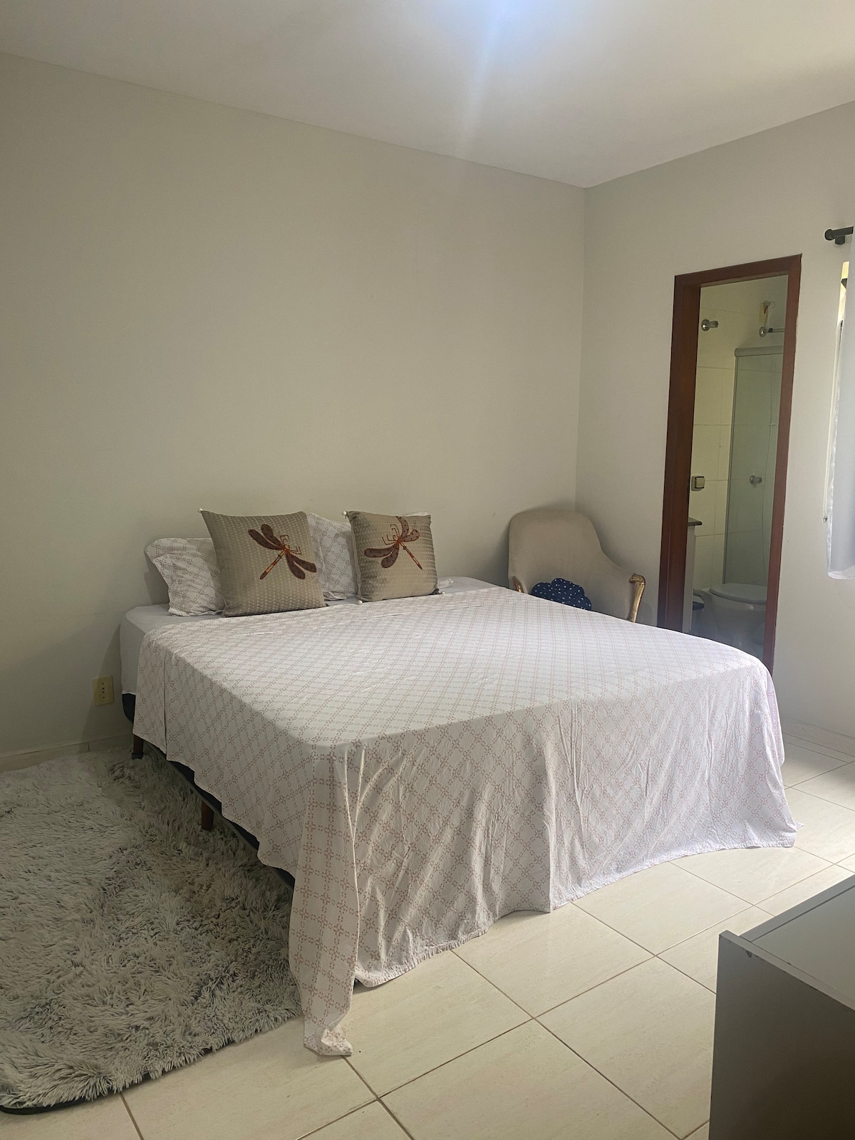 A cozy bedroom features a spacious bed covered with a light-colored blanket and two decorative pillows. A small rug lies on the floor, and a soft armchair is positioned nearby. A door leads to a bathroom, and natural light enters through a window.