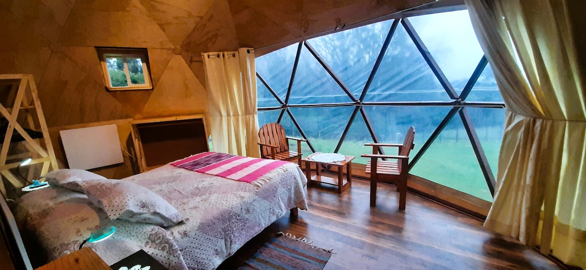 A cozy sleeping area features a spacious bed adorned with neutral linens and a striped blanket. Large triangular windows provide views of the surrounding landscape, while two wooden chairs and a small table complement the warm wooden flooring.