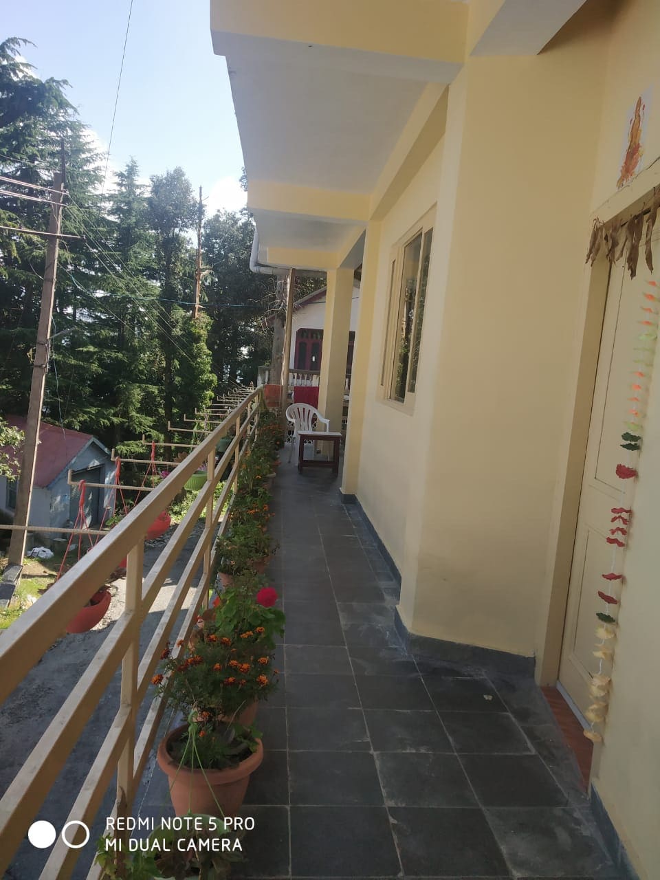 A covered balcony extends alongside the building, featuring a light-colored railing adorned with potted flowers. A white chair is positioned against the wall, inviting relaxation. Lush trees and distant rooftops are visible in the background, contributing to a tranquil outdoor space.