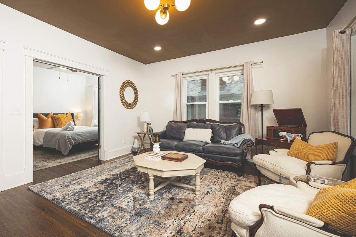 A cozy living room is highlighted by natural light streaming through a window. A dark leather sofa and two upholstered chairs face a central coffee table on an area rug. A wall-mounted mirror reflects the room's spaciousness, while a doorway leads to a separate bedroom.