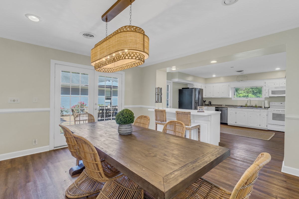A spacious dining area features a large wooden table surrounded by wicker chairs. A woven light fixture hangs above, providing ample illumination. The open layout leads into a bright kitchen, with views of the outdoor space visible through the glass doors.