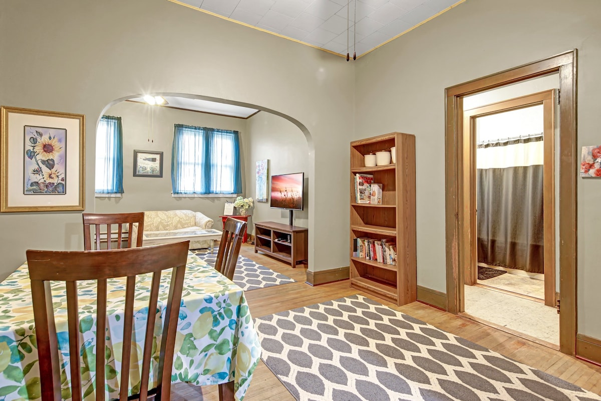 An open living area features a wooden dining table surrounded by four chairs and a patterned tablecloth. A futon is positioned against the wall with a television mounted nearby. A bookshelf filled with items is present, and a doorway leads to a bathroom area.