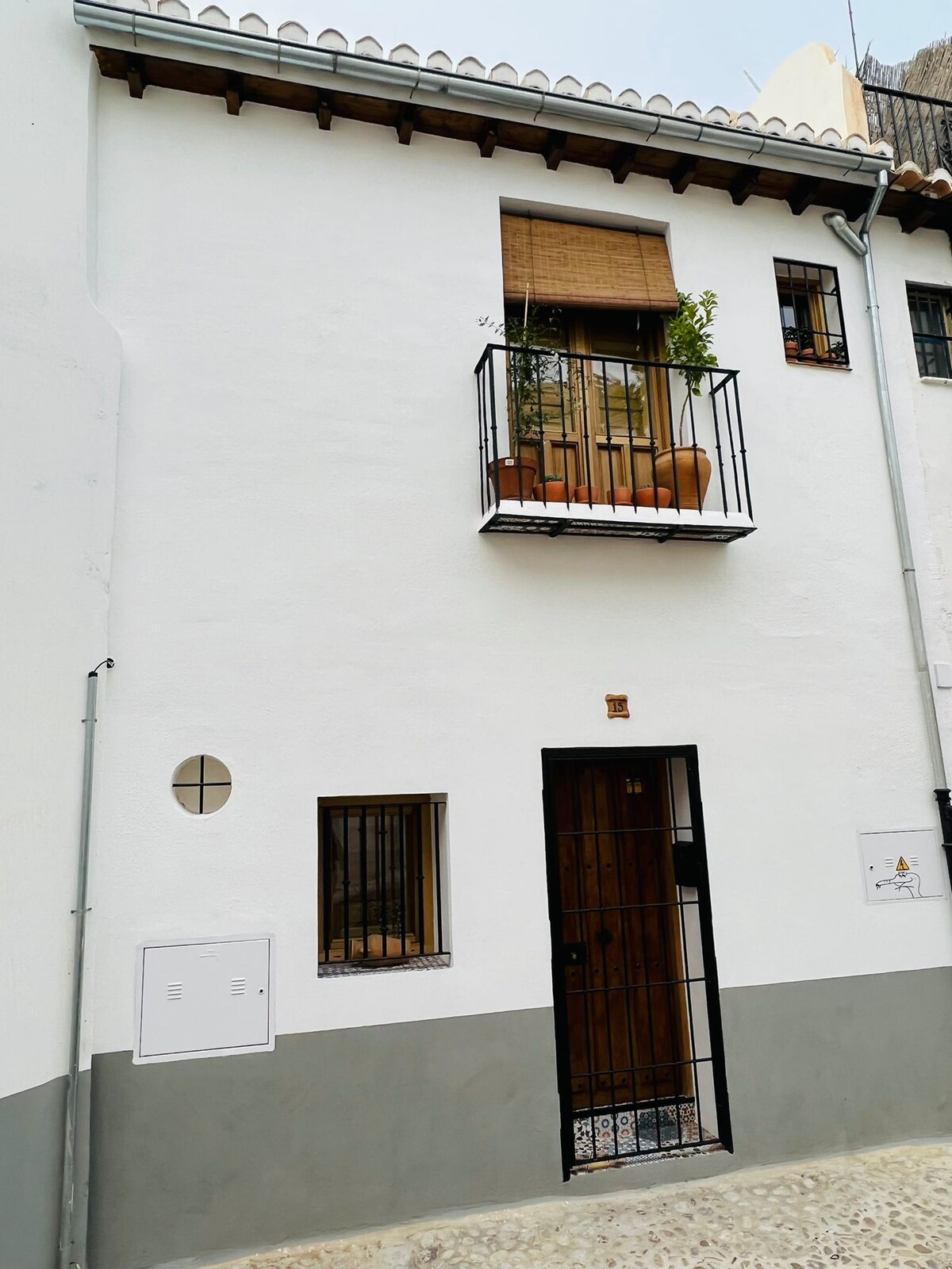 A charming exterior of a three-story house is presented, featuring a whitewashed façade with a decorative balcony adorned with potted plants. The entrance is framed by a simple wooden door and a black metal gate. Windows with dark frames contribute to the inviting appearance.