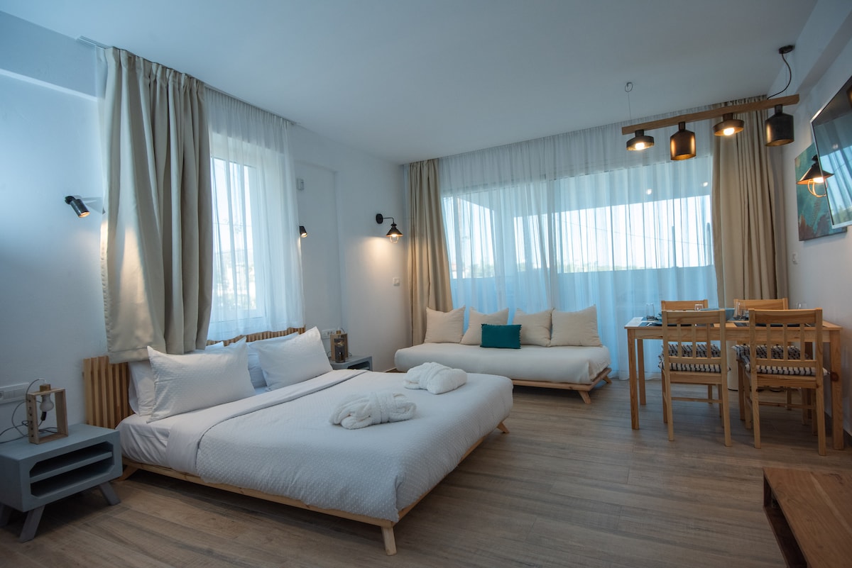 A bright and inviting studio features a spacious bed with white linens and a cozy daybed beside it. Natural light filters through sheer curtains, illuminating the wooden furnishings, a dining table, and soft accent pillows. Well-placed lighting provides a relaxing ambiance.