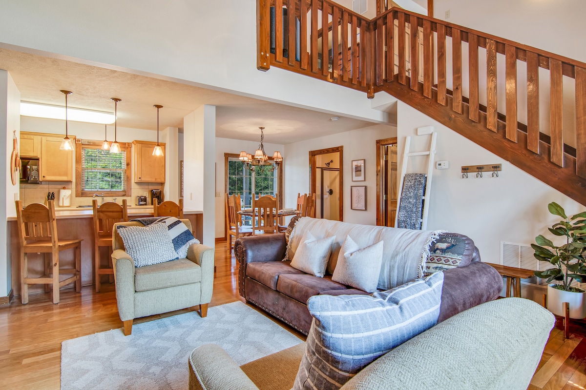 An inviting living area features a variety of seating options, including a brown sofa and a light green armchair. Warm lighting from pendant fixtures highlights the open layout connecting to the kitchen. A wooden staircase leads to the upper level, while large windows provide natural light.