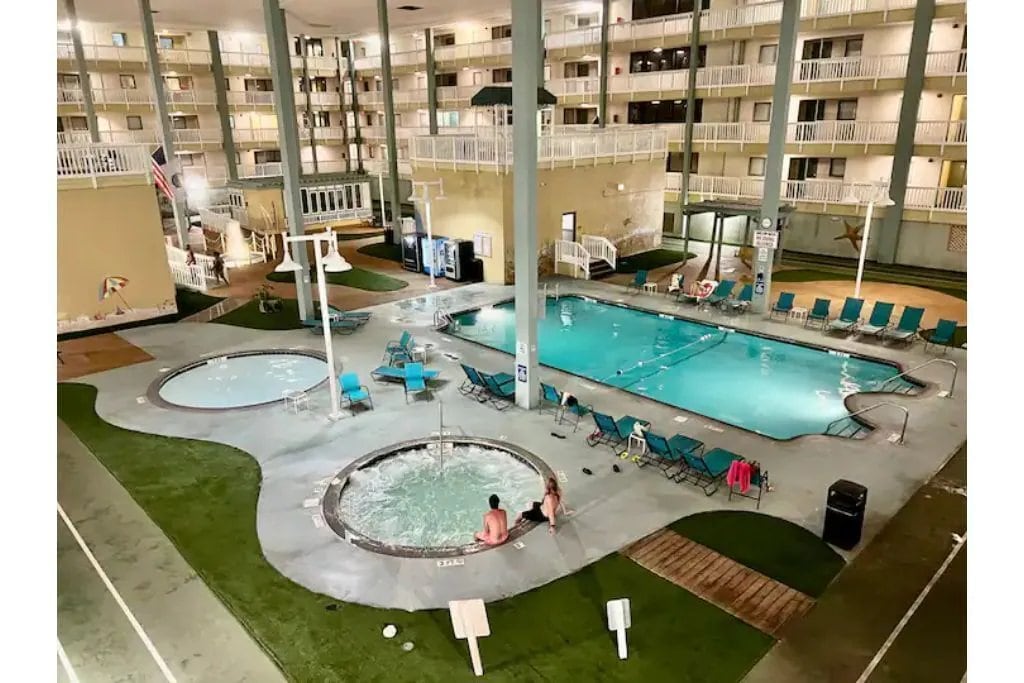 The image captures a spacious indoor pool area, featuring a large swimming pool, hot tub, and lounging chairs. Surrounding the pool, green turf enhances the leisure space. Multiple levels of the resort are visible in the background, showcasing a well-lit and inviting environment.