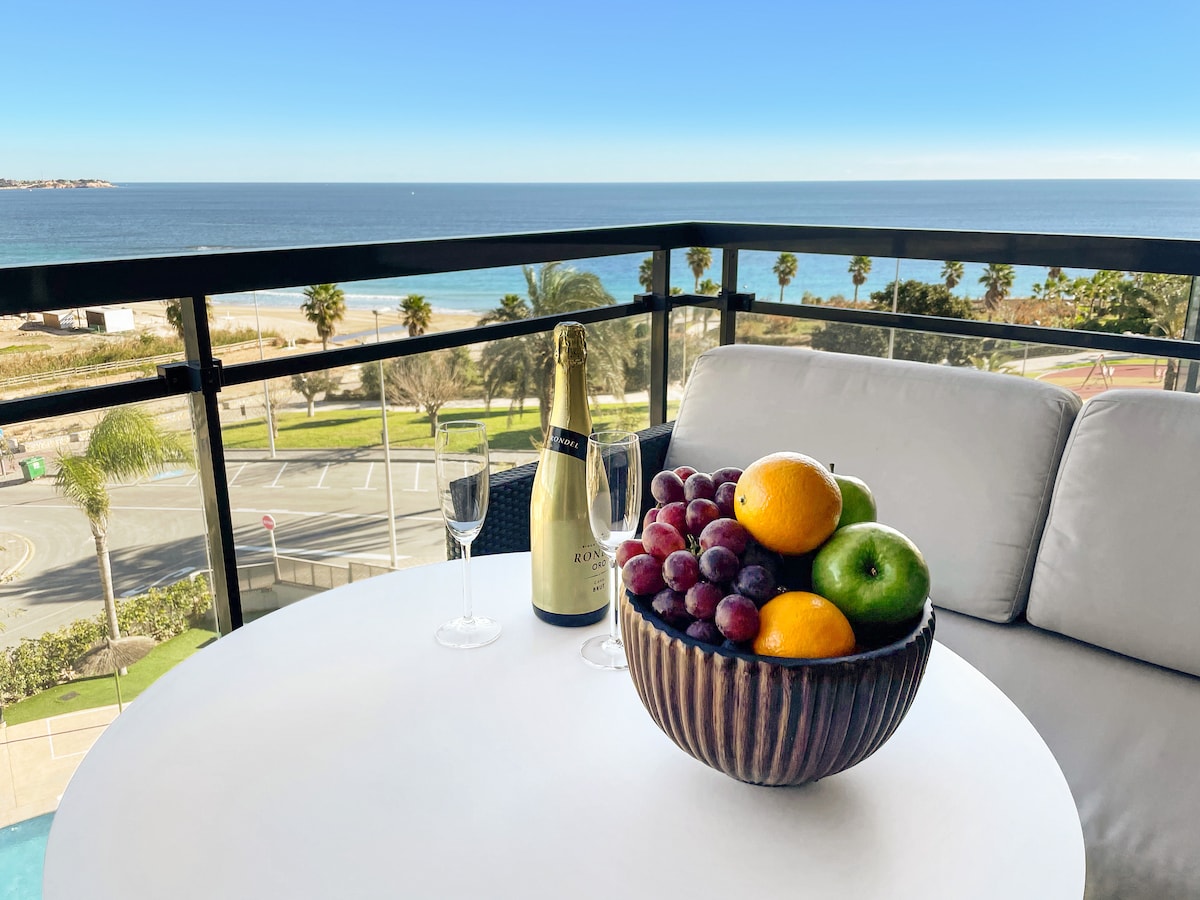 A round table is set on a balcony, featuring a bowl of fresh fruits including grapes, an orange, and a green apple. Beside the bowl are two fluted glasses and a bottle of champagne, with a clear view of the blue sea and palm trees in the background.