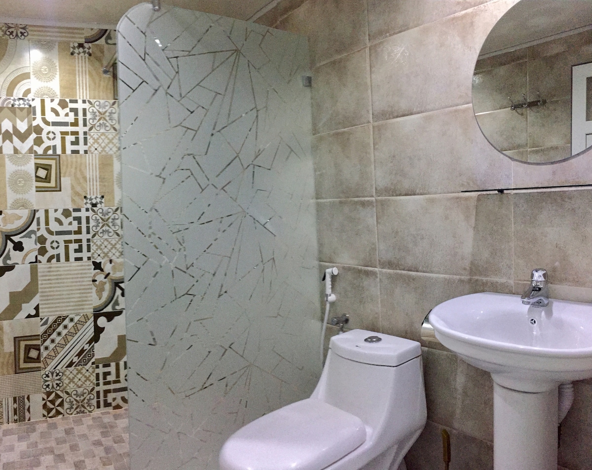 A modern bathroom features a clear glass shower enclosure beside a white toilet. A round mirror is mounted above a white pedestal sink, which complements the patterned wall tiles. The space is well-lit, emphasizing the clean and functional design.