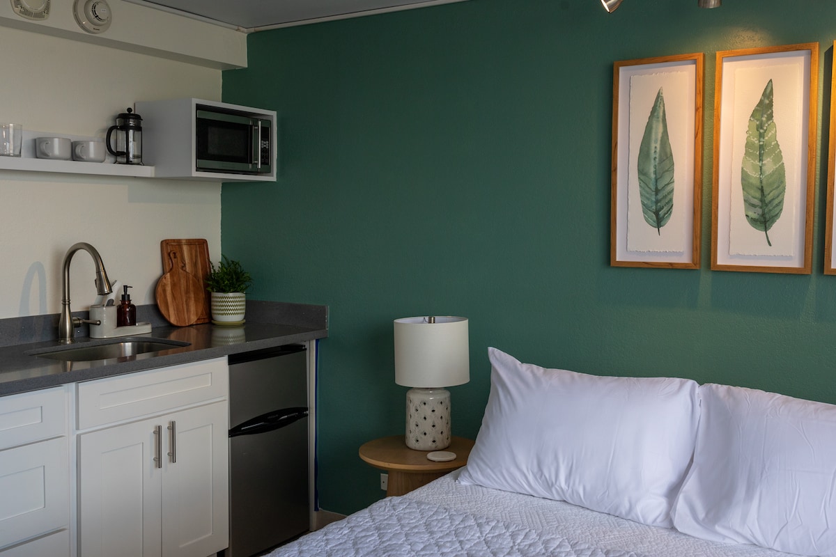A neatly arranged studio space features a spacious bed with white linens near a kitchenette. A small countertop is equipped with a microwave and coffee maker. Two framed leaf prints decorate the wall above the bed, complementing the green accent wall.