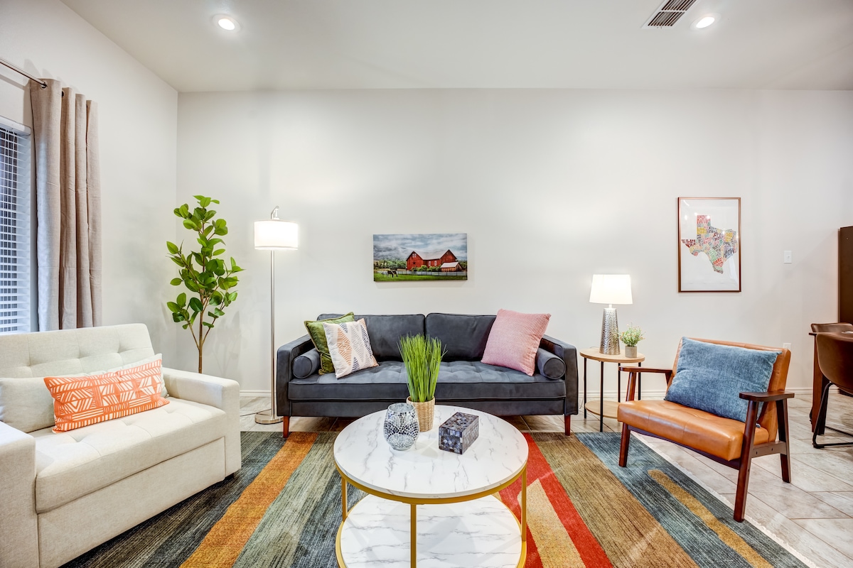 A bright living area features a comfortable, dark sofa and cozy chairs, complemented by colorful throw pillows. A marble coffee table sits at the center, surrounded by a vibrant area rug. Soft lighting from floor and table lamps enhances the welcoming environment.