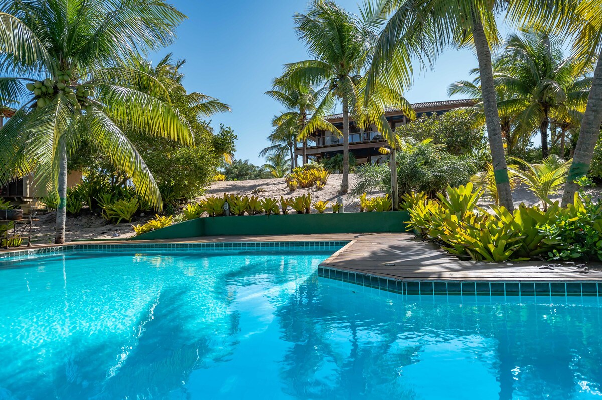 A clear swimming pool reflects the sunlight, surrounded by lush greenery and palm trees. The vibrant landscaping features various tropical plants along the pool's edge, enhancing the tranquil outdoor space. In the background, a multi-level house is partially visible, nestled among the vegetation.