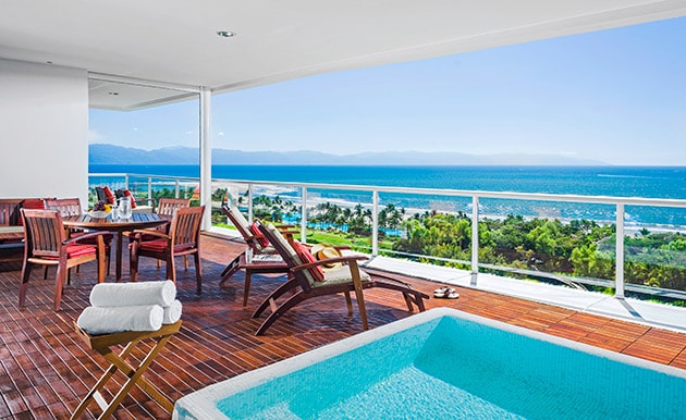 A spacious outdoor balcony features a plunge pool surrounded by wooden decking. Comfortable lounge chairs and a dining table invite relaxation while offering stunning views of the ocean and lush landscapes. Clear skies and distant mountains enhance the serene atmosphere.