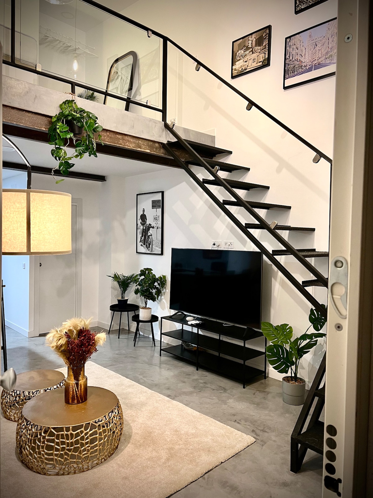 An open living area features a modern staircase leading to a lofted space. A large television is positioned on a sleek stand, surrounded by various indoor plants. A round coffee table with a textured surface sits on a light area rug, adding a cozy touch to the space.