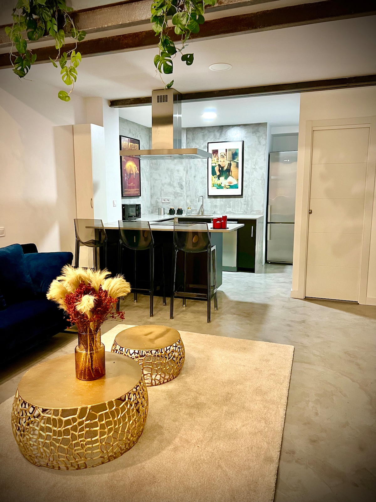 A modern loft showcases a stylish living area with a blue sofa and decorative gold tables. The kitchenette is visible in the background, featuring bar seating and elegant decor. Lush greenery is suspended from the ceiling, adding a touch of nature to the design.