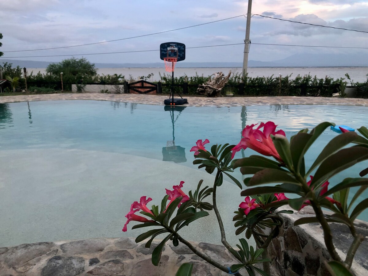 A serene swimming pool reflects the sky, bordered by lush greenery and colorful flowers. A basketball hoop is positioned by the water's edge, providing recreational opportunities. The distant horizon showcases a tranquil view of the water, enhancing the peaceful ambiance of the space.