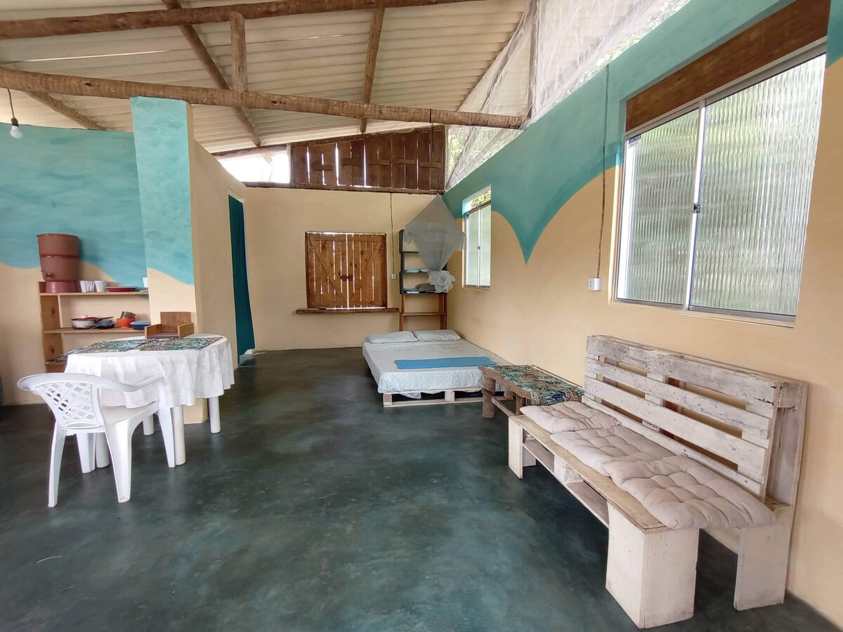 An open living area is highlighted by light blue walls and natural light from a large window. A simple wooden bench and a small table with chairs occupy the space. Two spacious mattresses are placed on the floor, offering a flexible sleeping arrangement.