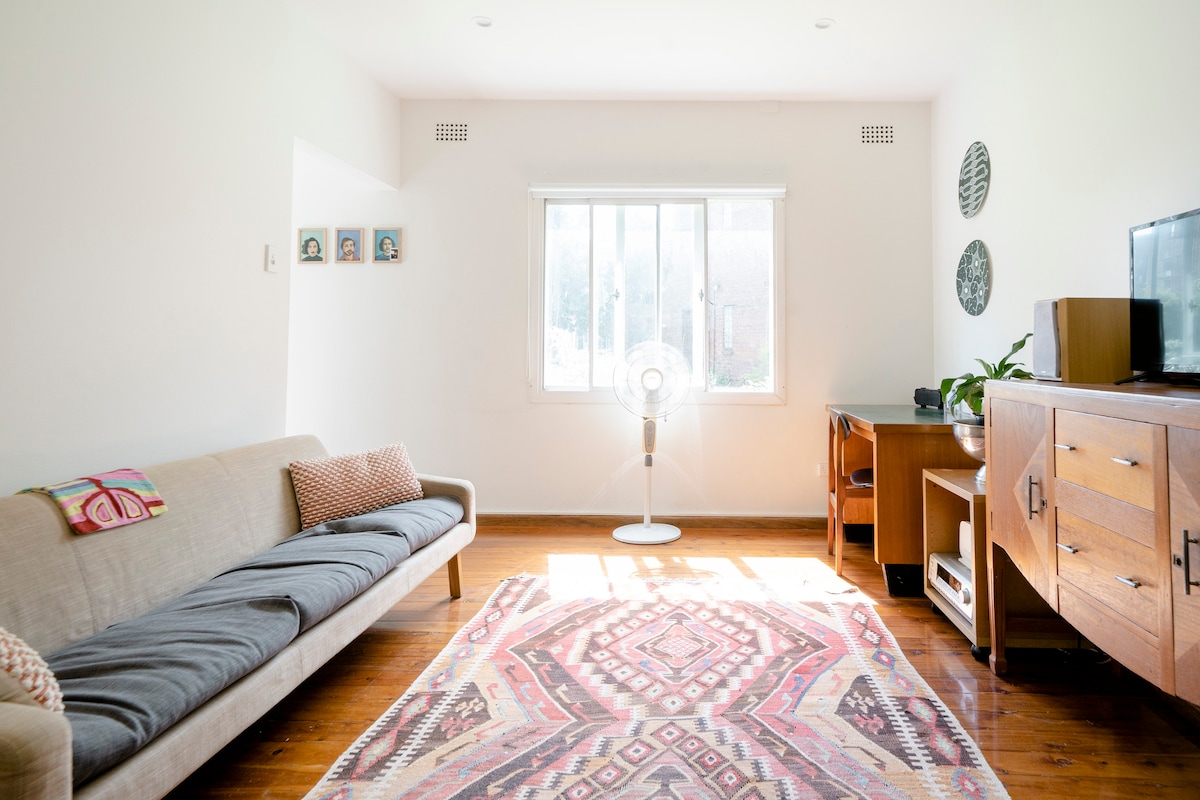 A clean living area features a light-colored sofa, complemented by decorative cushions. A patterned rug adds color to the wooden floor. A window allows natural light, and a fan stands beside it. A wooden cabinet and a television are also present.