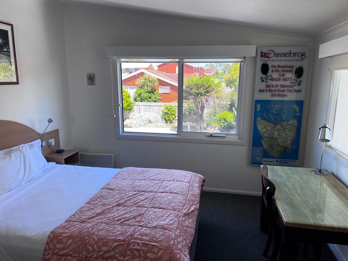 A comfortable room features a queen bed with a light pink coverlet. A window offers views of the landscape outside, while a small desk is positioned beside it. Natural light brightens the space, complemented by neutral walls and a simple accent decor.
